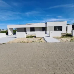 Nowoczesny, parterowy dom z płaskim dachem, częściowo wykończony, z widocznymi elementami stanu surowego, na pierwszym planie ułożona kostka brukowa.