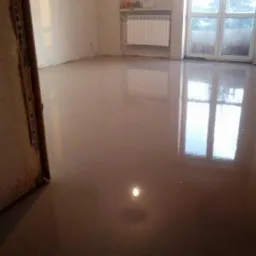 Wylany, mokry samopoziomujący podkład podłogowy w pokoju z grzejnikiem i oknem balkonowym, odbijającym się w gładkiej powierzchni.