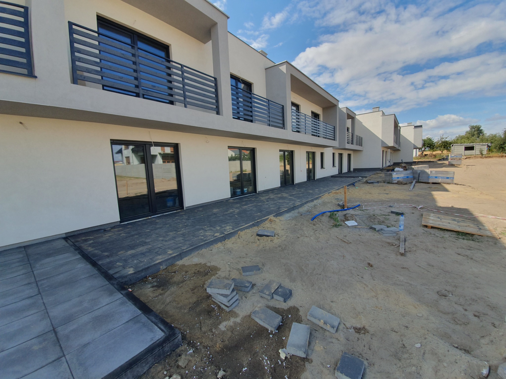 Nowoczesne domy w zabudowie szeregowej z ciemną kostką brukową na etapie układania, widoczne nieułożone kostki i piasek.