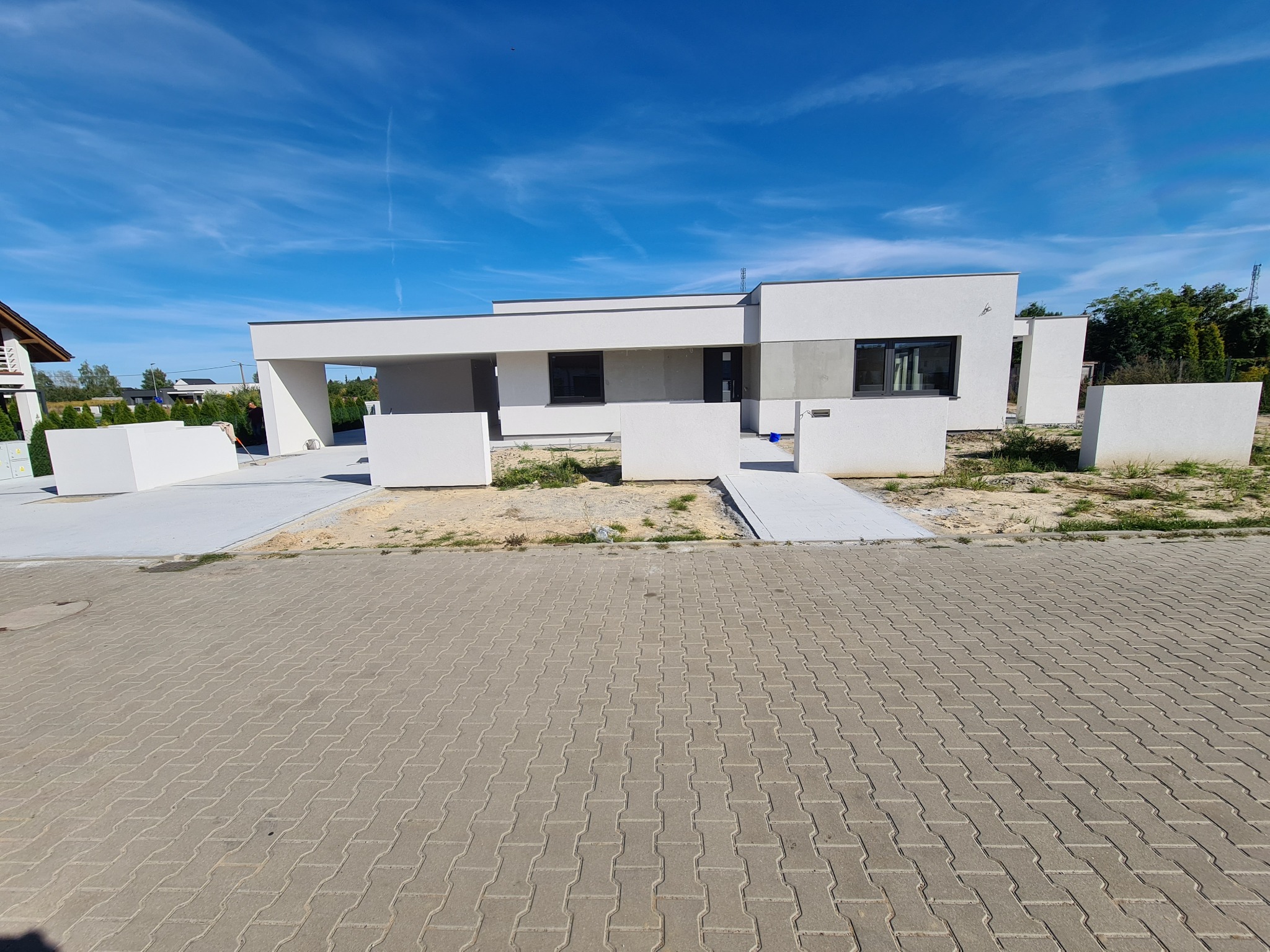 Nowoczesny, parterowy dom z płaskim dachem, częściowo wykończony, z widocznymi elementami stanu surowego, na pierwszym planie ułożona kostka brukowa.