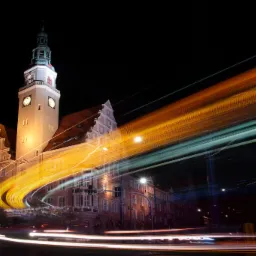 Nocne ujęcie oświetlonego budynku Ratusza Staromiejskiego w Gdańsku z długą ekspozycją świateł przejeżdżających pojazdów tworzących abstrakcyjne smugi.