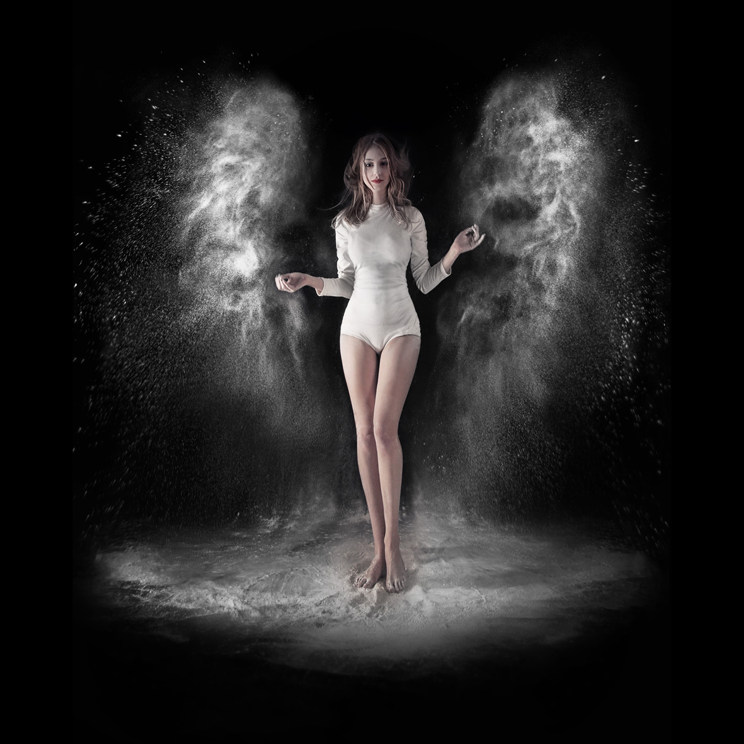 Kobieta w białym body, otoczona chmurami białego pyłu na czarnym tle, uchwycona w ruchu, z rozpuszczonymi włosami i delikatnym makijażem.