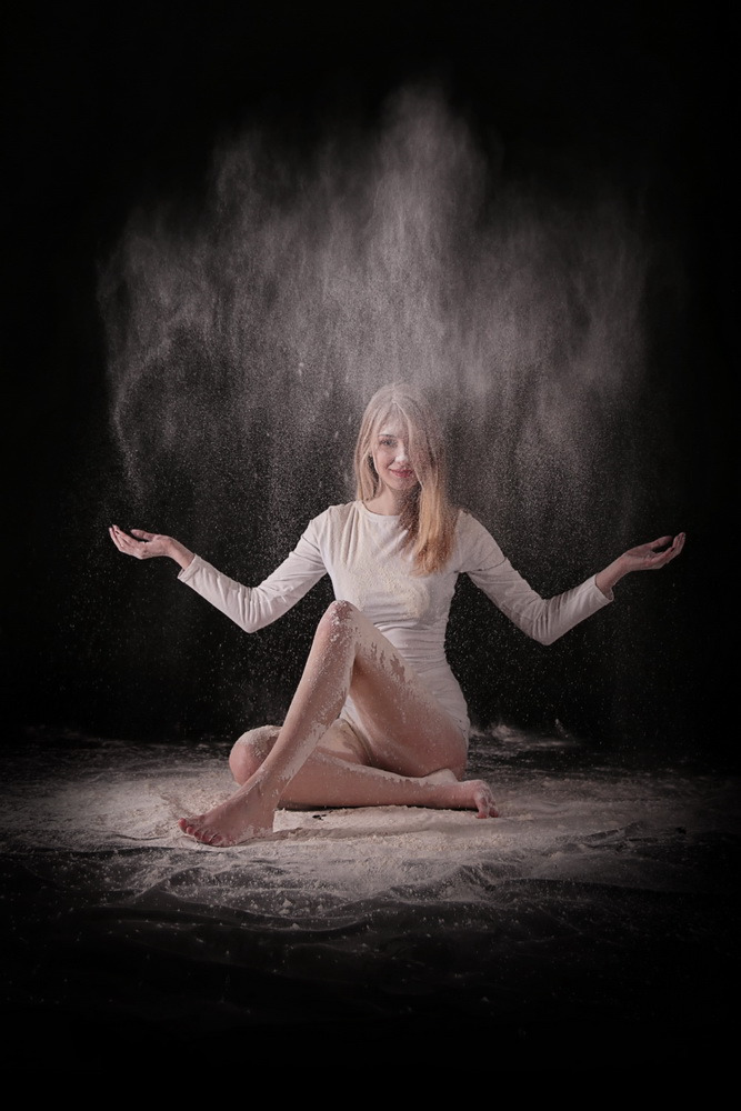 Kobieta w białym stroju siedzi na podłodze, na którą i na nią sypie się biały proszek na czarnym tle, oświetlona od góry.