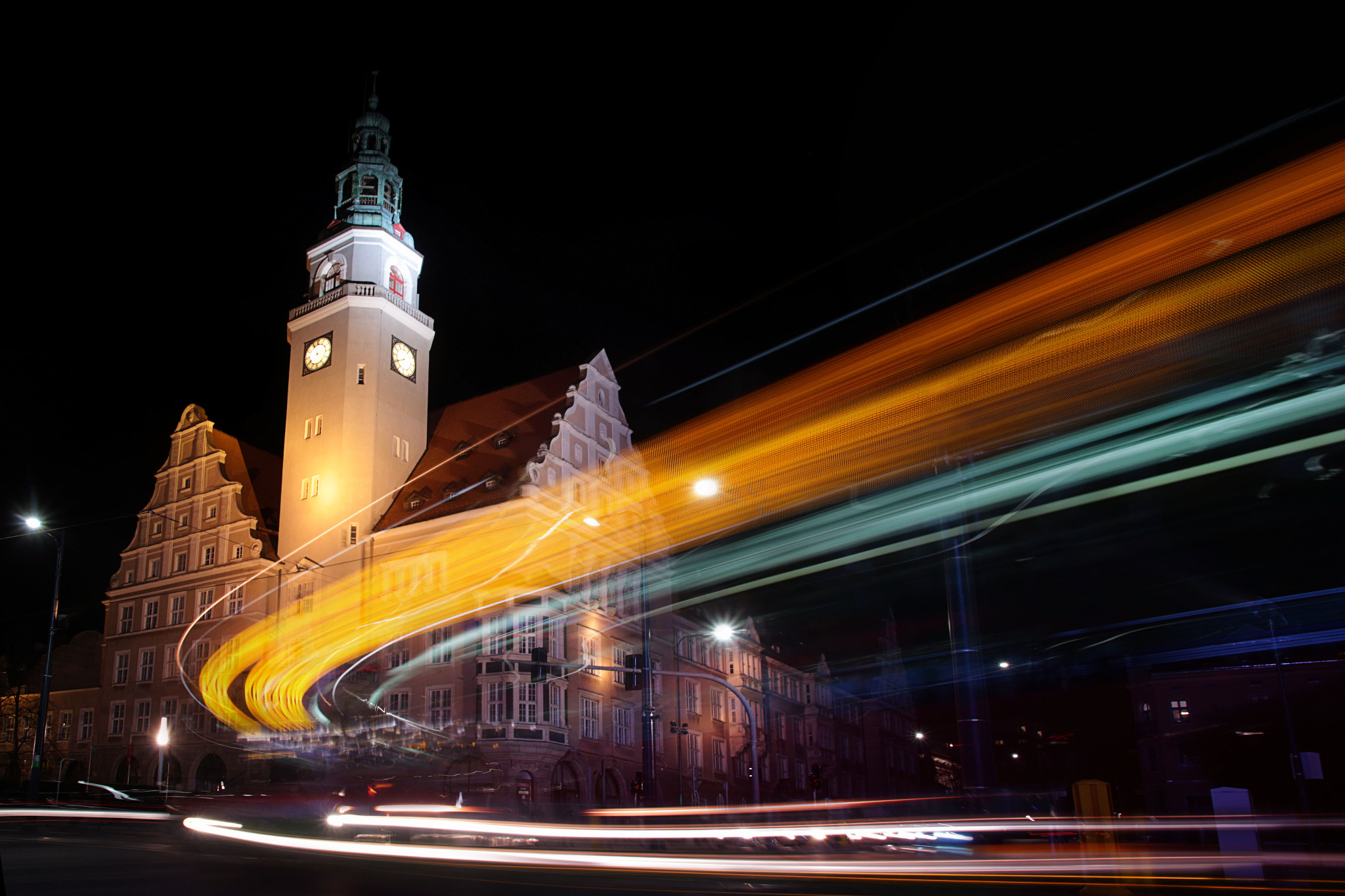 Nocne ujęcie oświetlonego budynku Ratusza Staromiejskiego w Gdańsku z długą ekspozycją świateł przejeżdżających pojazdów tworzących abstrakcyjne smugi.