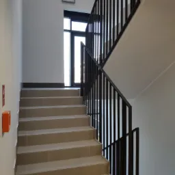 Klatka schodowa w nowoczesnym stylu z szarymi płytkami na stopniach, czarną metalową balustradą i białymi ścianami. Widoczny fragment okna na podeście. Na ścianie zamontowany pomarańczowy hydrant.