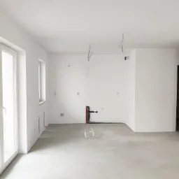Wnętrze mieszkania po malowaniu natryskowym na biało, widoczne przygotowane instalacje elektryczne i hydrauliczne, surowy betonowy podkład podłogowy.