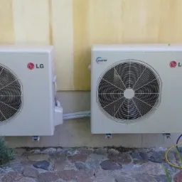 Dwa zamontowane na zewnątrz budynku klimatyzatory LG Inverter, połączone przewodami, widoczne na tle żółtej ściany i kamiennej podstawy z roślinnością.