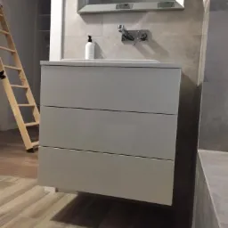 Szara szafka łazienkowa z trzema szufladami i umywalką nablatową, zamontowana na ścianie w Gościnie, z widoczną baterią i lustrem w ramie. W tle drabina oparta o ścianę.