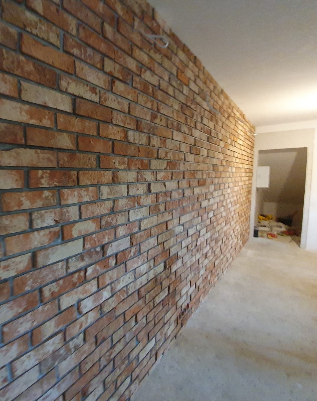 Ściana wyłożona cegłą klinkierową w odcieniach czerwieni i brązu, z szarymi fugami, widoczna z perspektywy bocznej w nowo wykończonym wnętrzu.