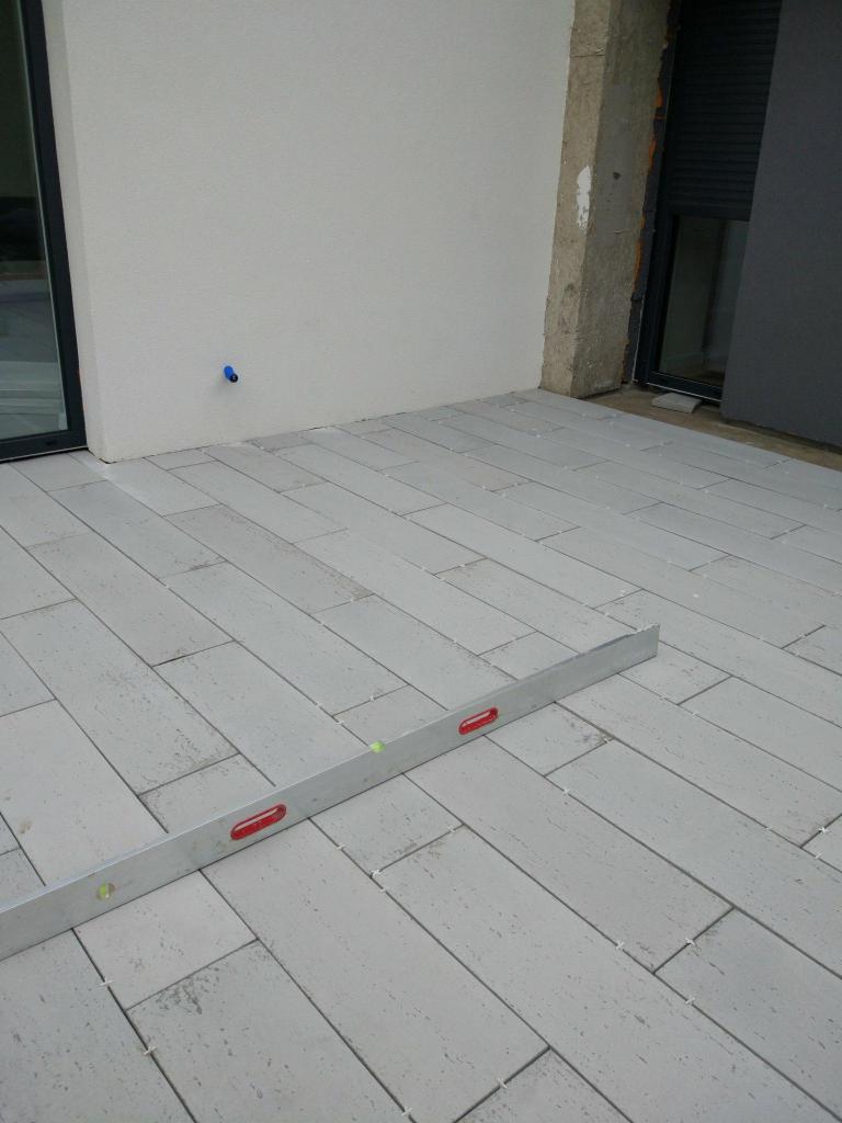 Ułożone szare płytki na podłodze balkonu, poziomica aluminiowa leży na płytkach, widoczne krzyżyki dystansowe między płytkami, ściana otynkowana na gładko z niebieskim zaworem