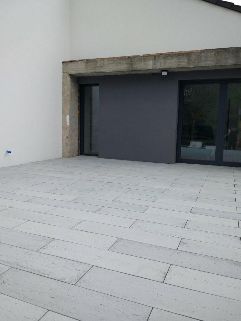 Wyłożony jasnoszarymi płytami taras przed budynkiem z ciemnoszarą ścianą i betonowym nadprożem, widoczne szklane drzwi przesuwne.