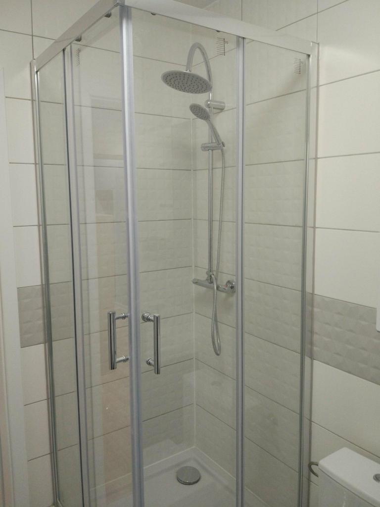 Kabina prysznicowa z deszczownicą i baterią termostatyczną, wykończona płytkami w odcieniach szarości i bieli z subtelną strukturą, widoczny fragment toalety.