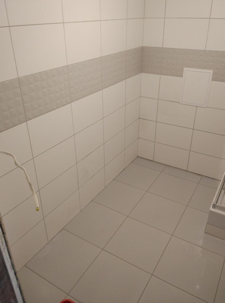 Świeżo wykafelkowana ściana i podłoga w łazience z dekoracyjnym pasem płytek z wypukłym wzorem, widoczny fragment kabla elektrycznego i klapa rewizyjna.