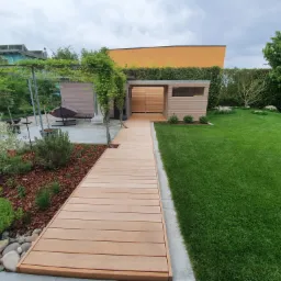 Nowoczesny ogród z drewnianą ścieżką prowadzącą do drewnianej altany z przesuwnymi drzwiami, trawnikiem i roślinnością.