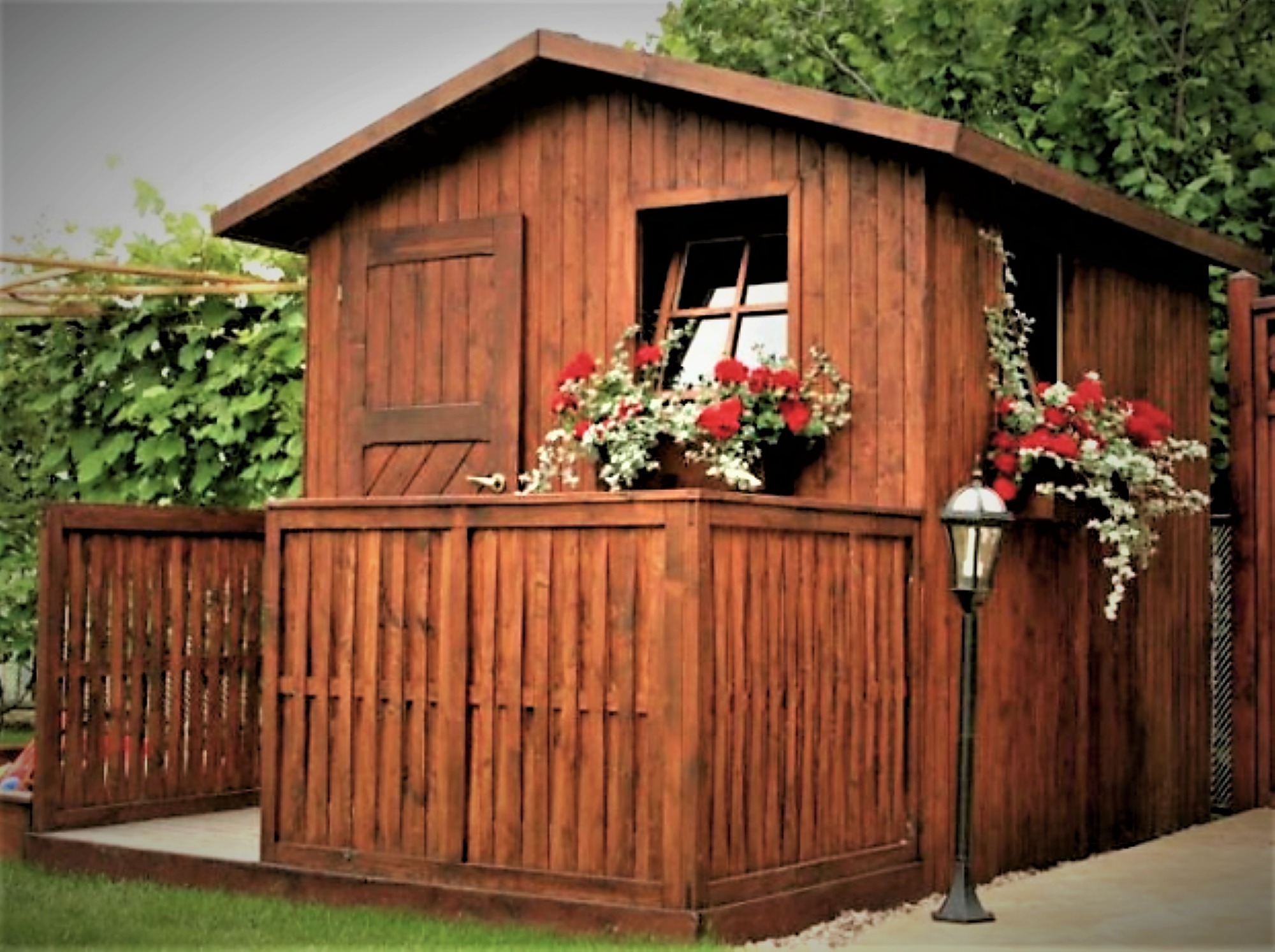 Drewniana konstrukcja z oknem i drzwiami, otoczona niskim płotkiem, udekorowana czerwonymi kwiatami w donicach, z lampą ogrodową obok, na tle zieleni.