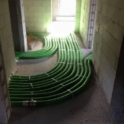 Wnętrze w trakcie budowy z ułożonymi zielonymi, karbowanymi rurami wentylacyjnymi, zabezpieczonymi metalowymi opaskami, prowadzącymi w kierunku jasnego otworu drzwiowego.