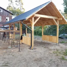 Drewniana wiata z ciemnym dachem i rynnami, częściowo zabudowana deskami, widoczna na tle brązowego domu i zieleni, z elementami rusztowania z przodu.