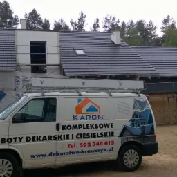 Nowy dach z ciemnej dachówki na domu w trakcie budowy, widoczny samochód firmy dekarskiej z reklamą.