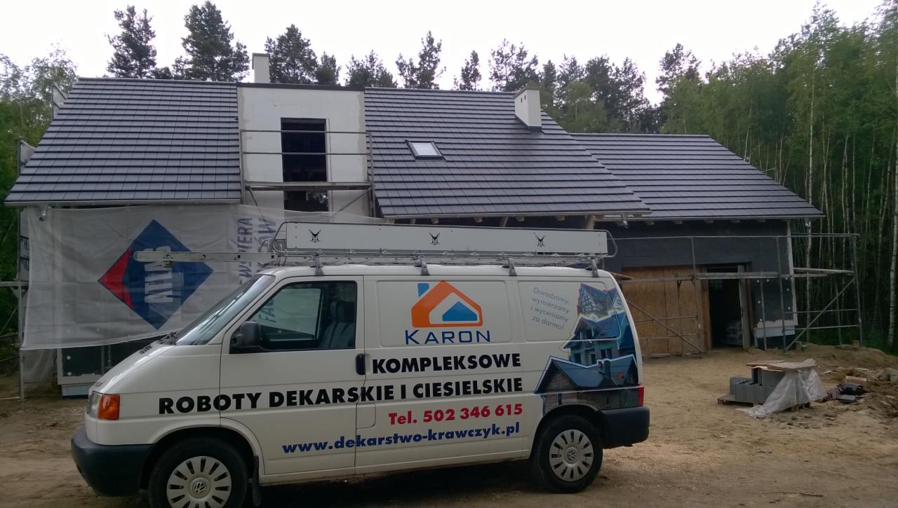 Nowy dach z ciemnej dachówki na domu w trakcie budowy, widoczny samochód firmy dekarskiej z reklamą.