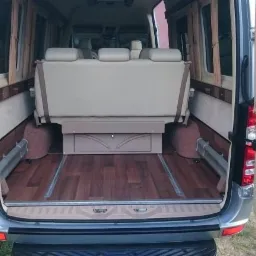 Wnętrze luksusowego busa z rozłożonymi siedzeniami, drewnianą podłogą i zasłonami w oknach, widok z otwartych tylnych drzwi.