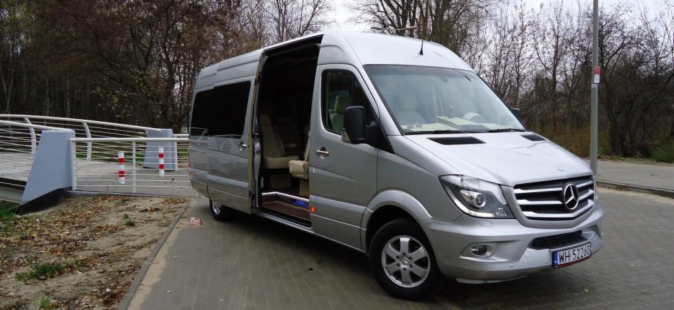 Srebrny van z otwartymi drzwiami przesuwnymi, widoczne wnętrze z jasną tapicerką, na tle drzew i barierki.