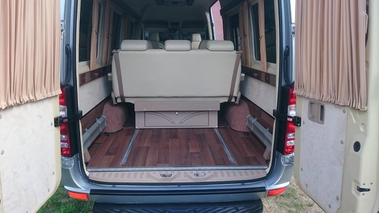 Wnętrze luksusowego busa z rozłożonymi siedzeniami, drewnianą podłogą i zasłonami w oknach, widok z otwartych tylnych drzwi.