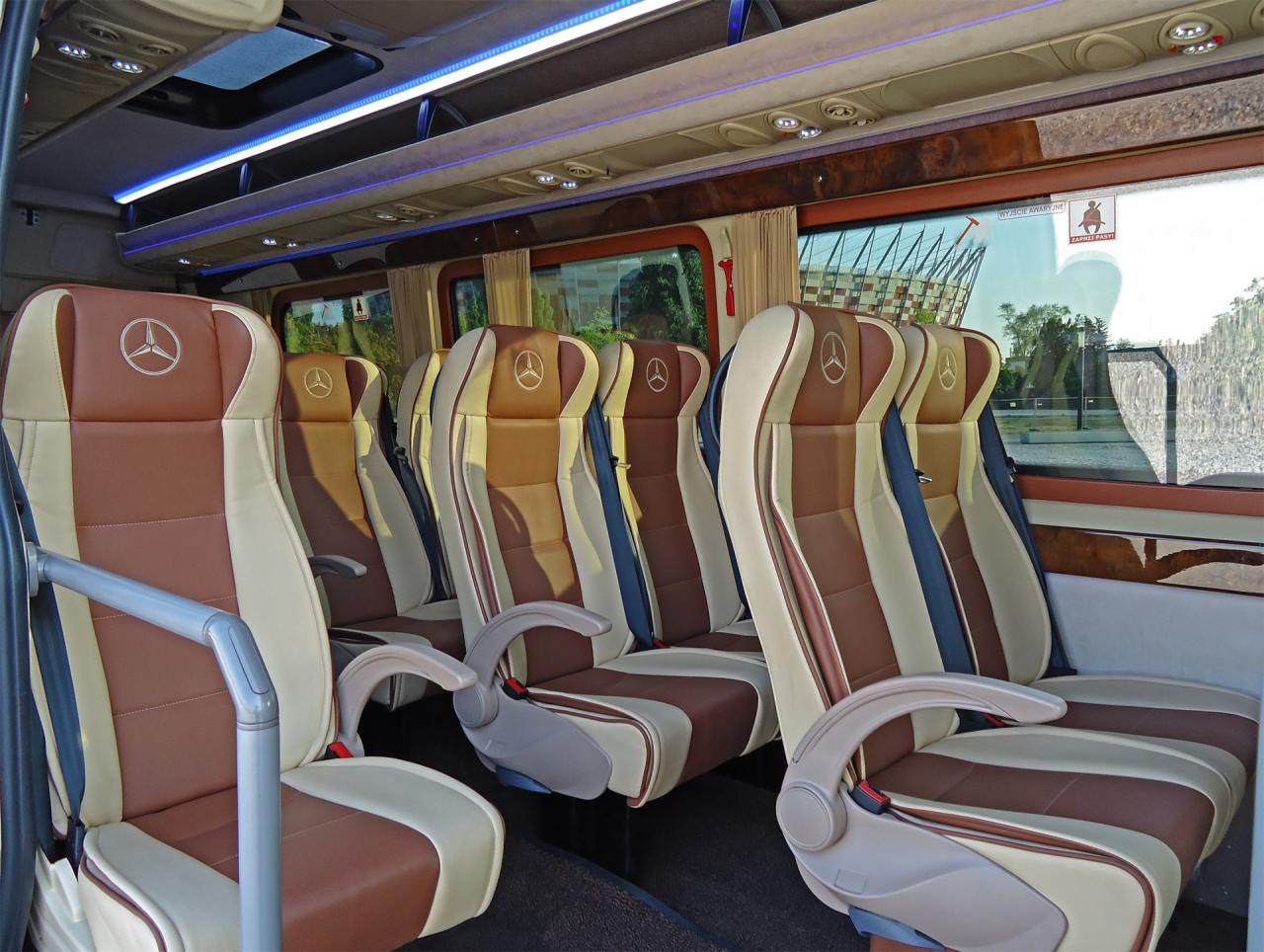Wnętrze luksusowego busa marki Mercedes z beżowo-brązowymi skórzanymi fotelami z logo Mercedes na zagłówkach, widok na okno z widocznym stadionem w tle.