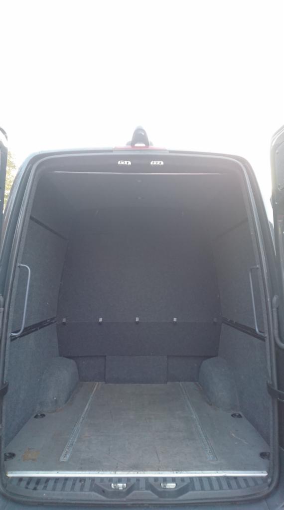 Wnętrze furgonetki z otwartymi tylnymi drzwiami, wyłożone szarą wykładziną, z metalowymi prowadnicami na podłodze.