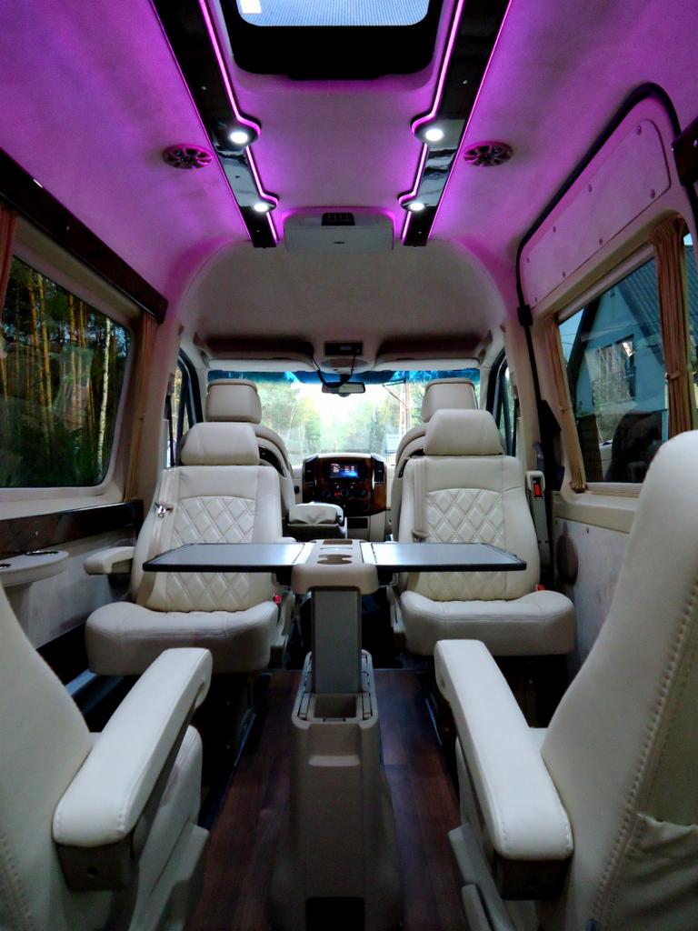Luksusowe wnętrze busa z beżowymi skórzanymi fotelami, rozkładanymi stolikami i fioletowym oświetleniem LED podsufitki.