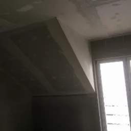 Skośny sufit poddasza w trakcie prac wykończeniowych, widoczne łączenia płyt gipsowo-kartonowych i wstępne szpachlowanie, okno w tle.