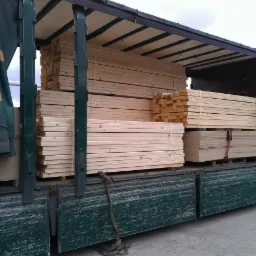 Ciężarówka z odkrytą naczepą załadowana drewnem budowlanym i płytami drewnopochodnymi, zabezpieczonymi pasami transportowymi.