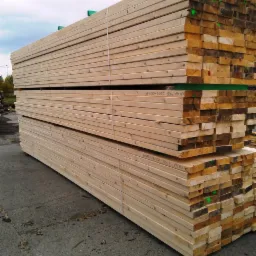 Stos tarcicy budowlanej, różne długości, spięty białym sznurkiem, widoczne oznaczenia wymiarów i nazwy producenta na niektórych deskach.