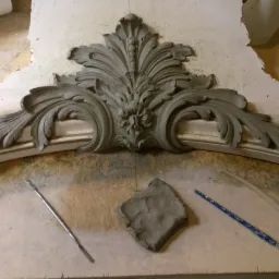 Fragment gipsowej dekoracji architektonicznej w stylu rokoko, leżący na drewnianym blacie, obok narzędzi modelarskich i bryłki gliny.