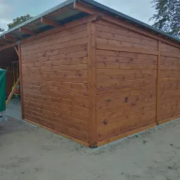 Zewnętrzny widok na nowo wybudowaną, drewnianą konstrukcję z płaskim dachem, prawdopodobnie altanę lub domek narzędziowy, z widocznymi śladami świeżego lakieru na brązowych deskach i szarą blachą...