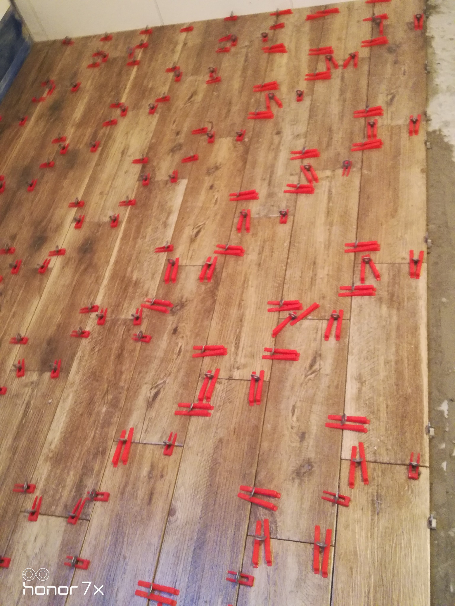 Układanie płytek imitujących drewno z użyciem czerwonych klipsów poziomujących na podłodze, widoczny fragment ściany z pozostałościami zaprawy.