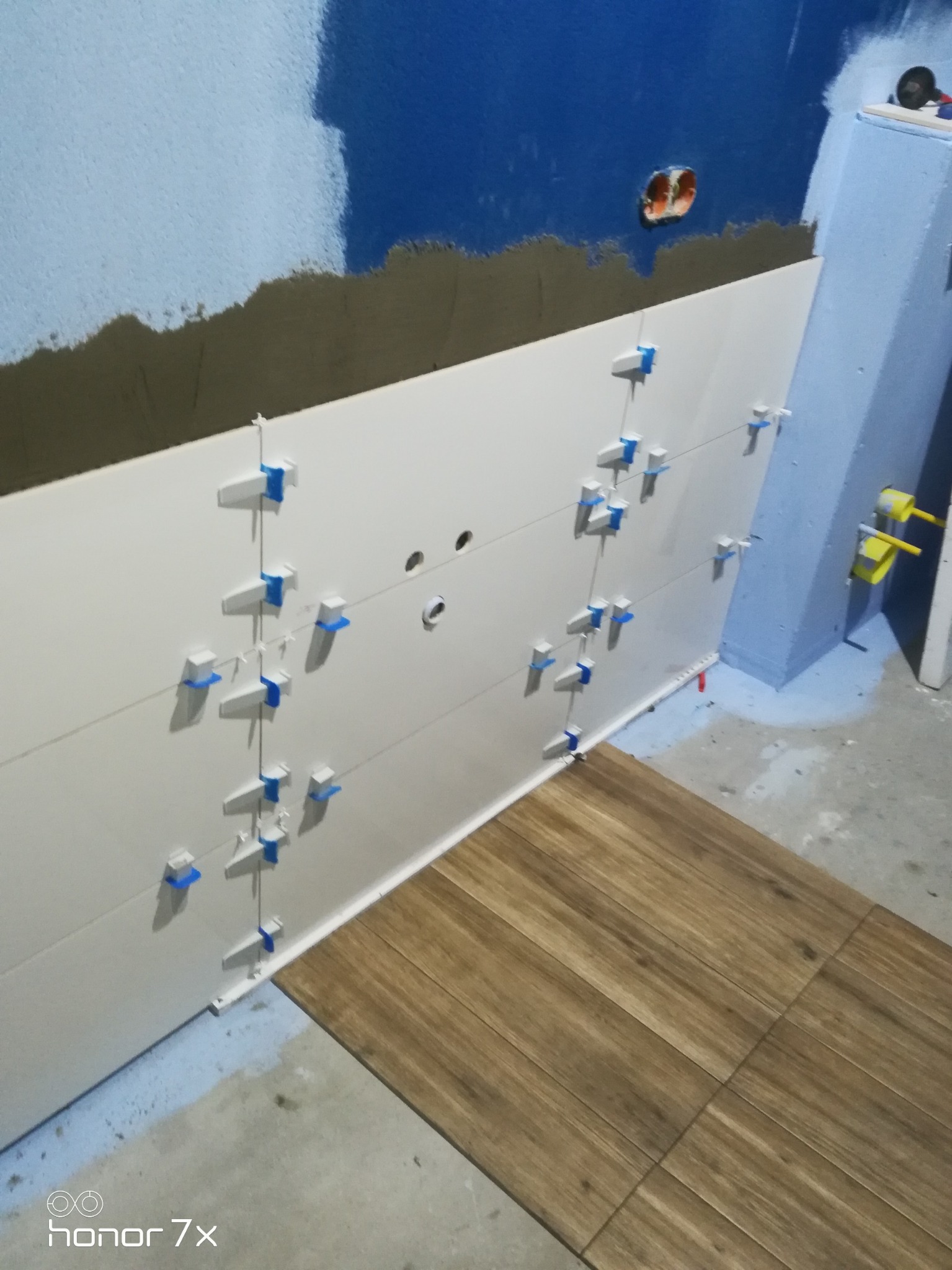 Ściana w trakcie układania białych płytek z systemem poziomowania, widoczne fragmenty podłogi z brązowych płytek imitujących drewno, oraz przygotowana ściana do dalszych prac.