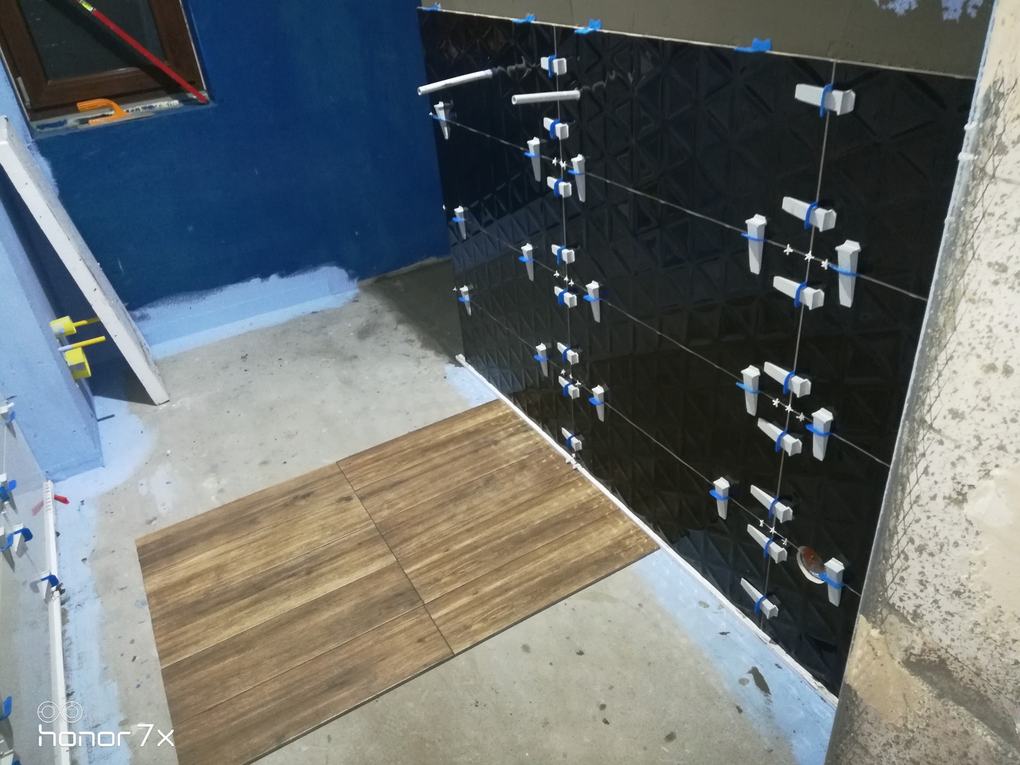 Układanie czarnych, wzorzystych płytek na ścianie z użyciem poziomicy i krzyżyków dystansowych; na podłodze ułożona część drewnopodobnych płytek.