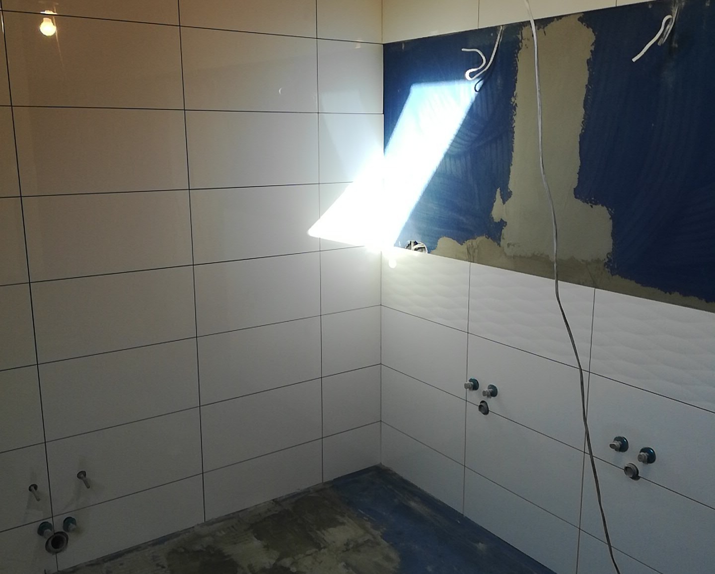 Wnętrze łazienki w trakcie remontu, z częściowo ułożonymi białymi, prostokątnymi płytkami na ścianach, widoczne wystające kable i instalacja wodna, ślady po tynku na górnej części ściany, podłoga...