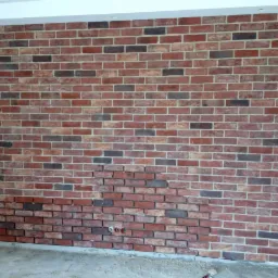 Częściowo wykończona ściana z cegły dekoracyjnej w odcieniach czerwieni i brązu, widoczne przewody elektryczne, fragment drabiny malarskiej w lewym dolnym rogu.