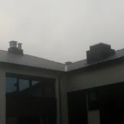 Nowoczesny dom z ciemną, metalową dachówką i kominami w pochmurny dzień.