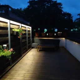 Wieczorny widok na oświetlony drewniany taras z drewnianą zabudową, na której wiszą donice z kwiatami, w tle widoczne drzewa i dekoracyjne oświetlenie.