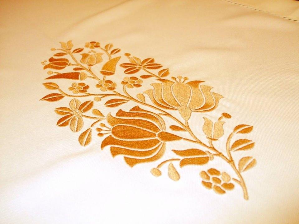Delikatny, złoty haft kwiatowy na kremowej tkaninie, przedstawiający stylizowane róże i inne kwiaty w owalnym układzie.