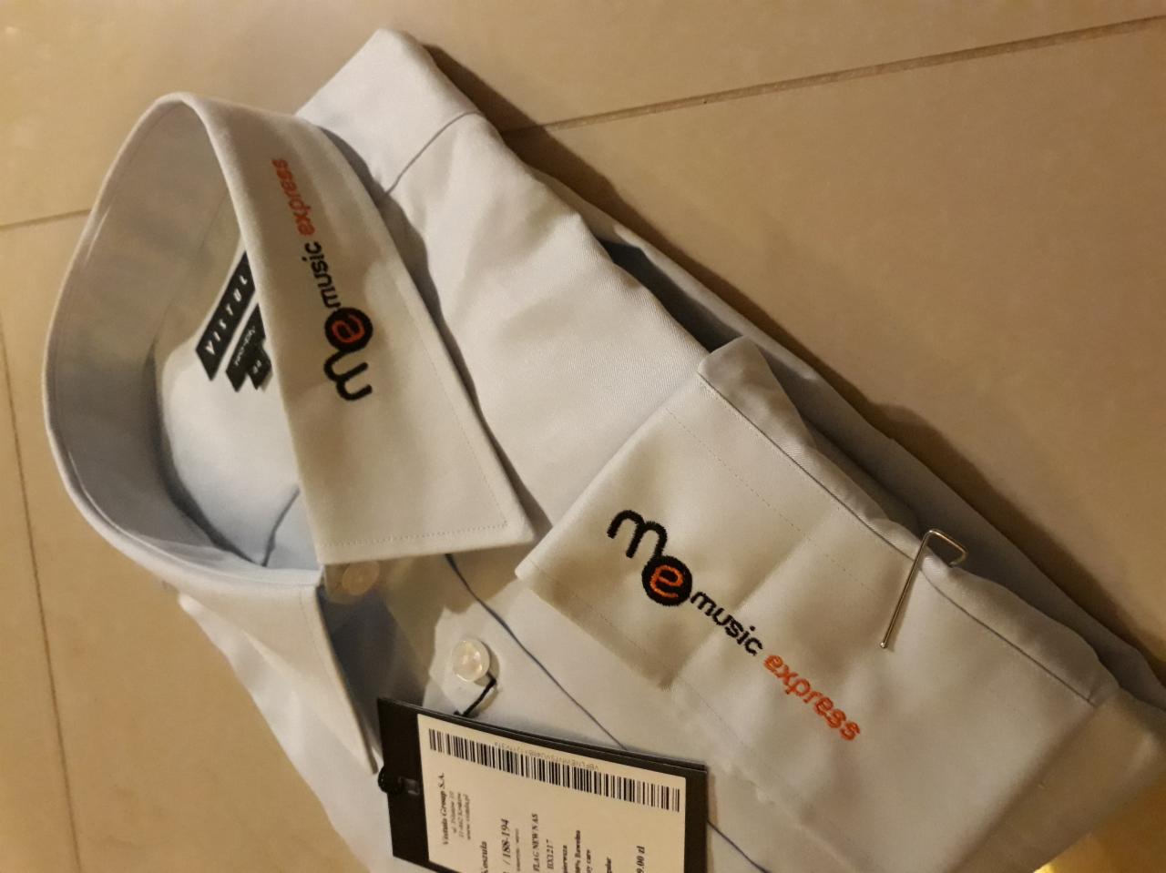 Jasnoniebieska koszula z wyhaftowanym logo 'me music express' na kołnierzyku i mankiecie, widoczna metka z ceną i szpilka.