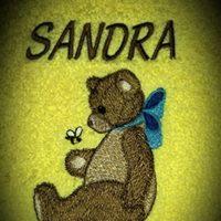 Żółta tkanina z wyhaftowanym imieniem 'SANDRA' nad brązowym misiem z niebieską kokardką, obok którego leci pszczoła.