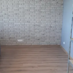 Ściana wyłożona dekoracyjną cegłą w odcieniach szarości, uzupełniona panelami podłogowymi w Ełku. Widoczne gniazdko elektryczne i fragment metalowego regału.