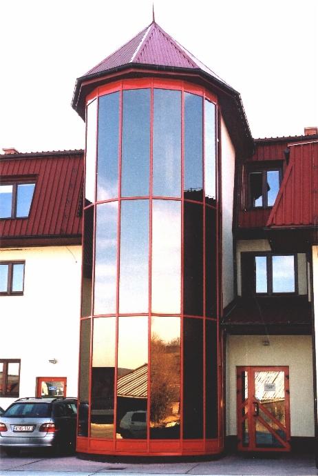 Szklana wieża z czerwonymi ramami na fasadzie budynku, odbijająca krajobraz, z samochodem zaparkowanym przed budynkiem.