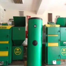 Zielone kotły grzewcze z żółtymi akcentami i logo KBO, ustawione w rzędzie w pomieszczeniu o jasnych ścianach, z widocznymi grzejnikami w tle.