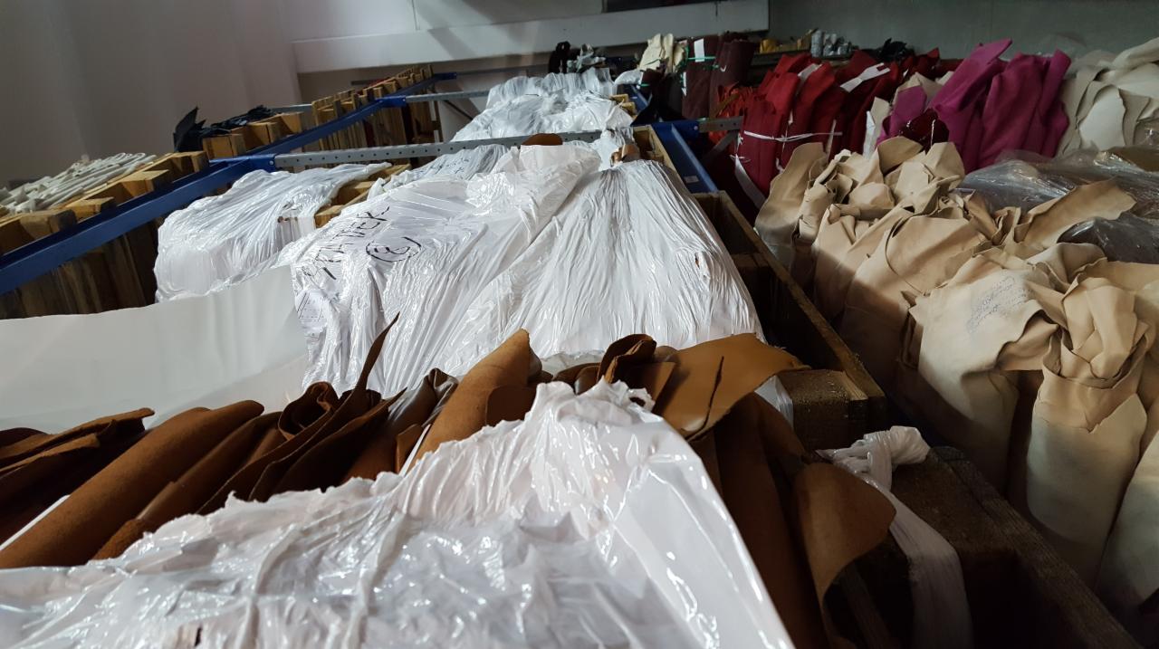 Magazyn skórzanych materiałów w rolach i pociętych arkuszach, częściowo zapakowanych w folię, ułożonych na drewnianych paletach i metalowych regałach, widoczne różne odcienie brązu, czerwieni i bieli.