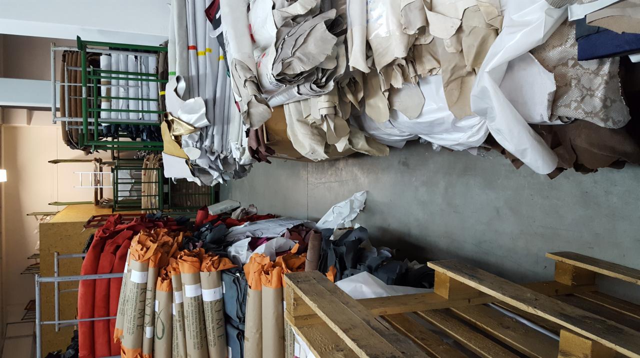 Magazyn z rolkami i pociętymi kawałkami różnokolorowej skóry i materiałów tapicerskich, ułożonych na metalowych regałach i drewnianych paletach.