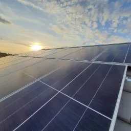 Panele słoneczne na dachu domu oświetlone zachodzącym słońcem, widoczne chmury na niebie.